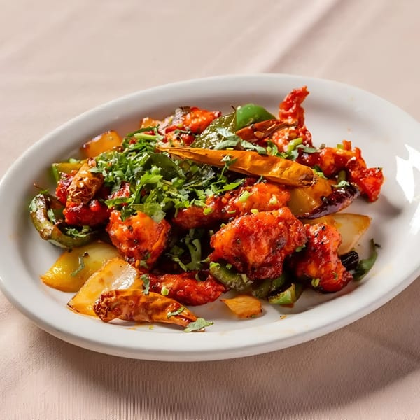 A white oval plate filled with bright red Indian chili chicken, green peppers, and cilantro.