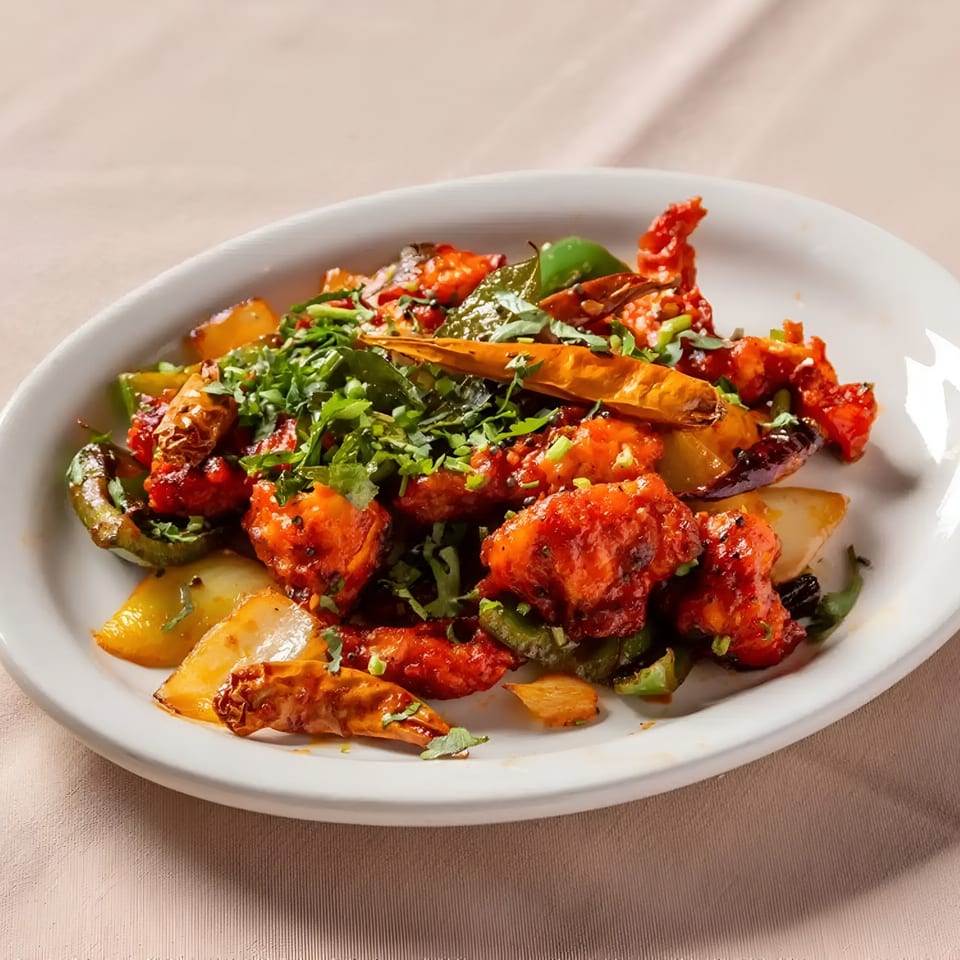 A white oval plate filled with bright red Indian chili chicken, green peppers, and cilantro.