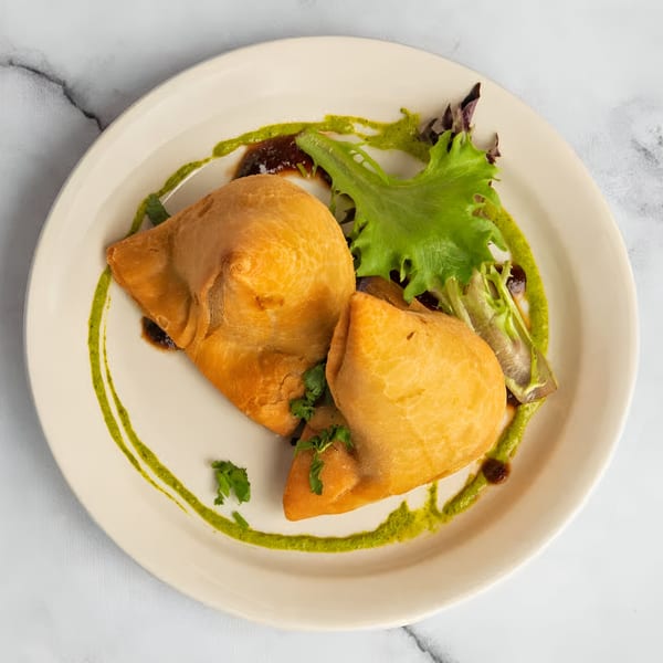 Two golden-brown samosas with green chutney, dark sauce, and fresh greens on a white plate.