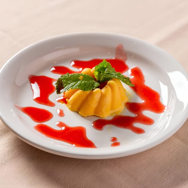 A yellow Indian dessert, possibly mango kulfi, garnished with mint and strawberry sauce on a white plate.